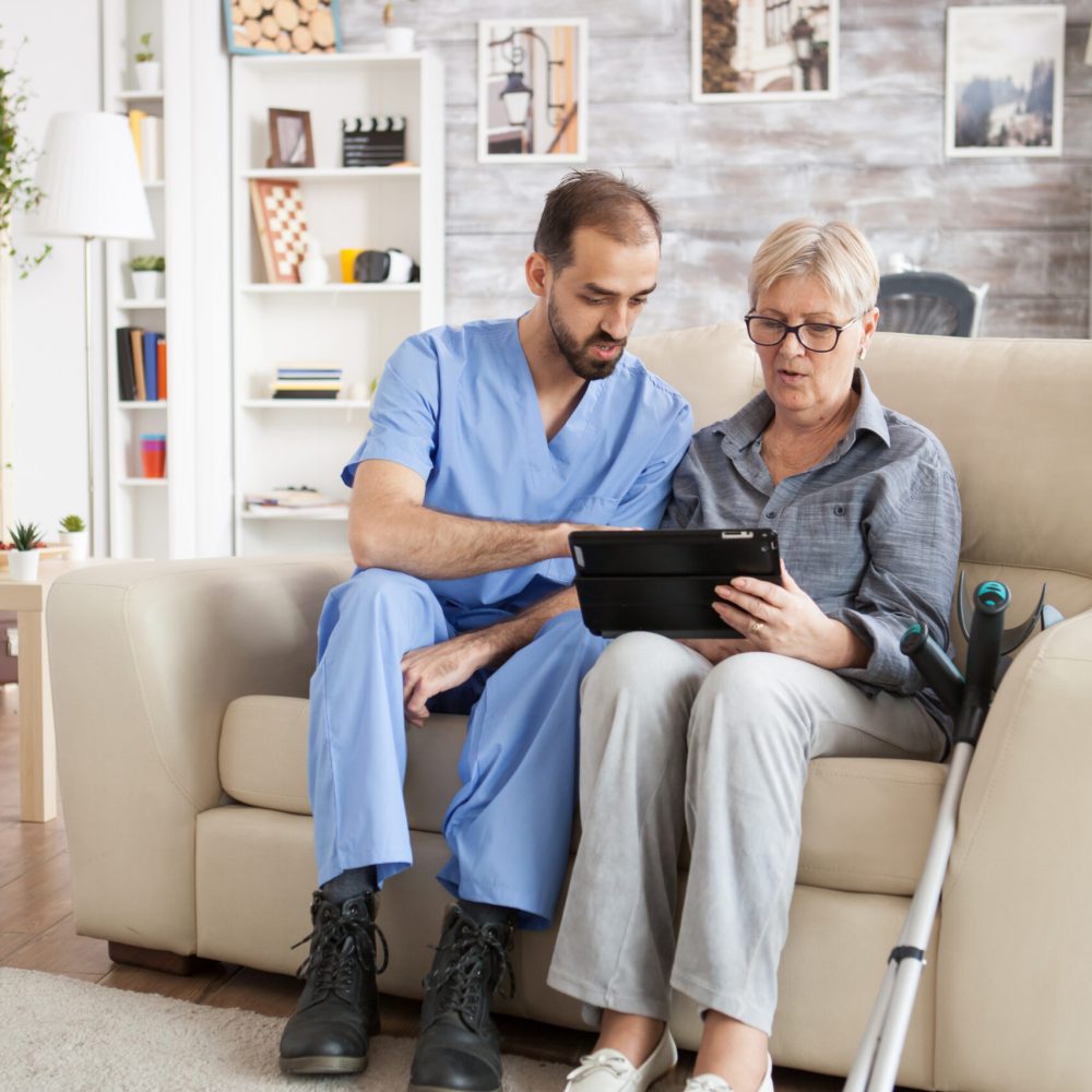 Young doctor in nursing home helping senior woman with crutches to use tablet computer.